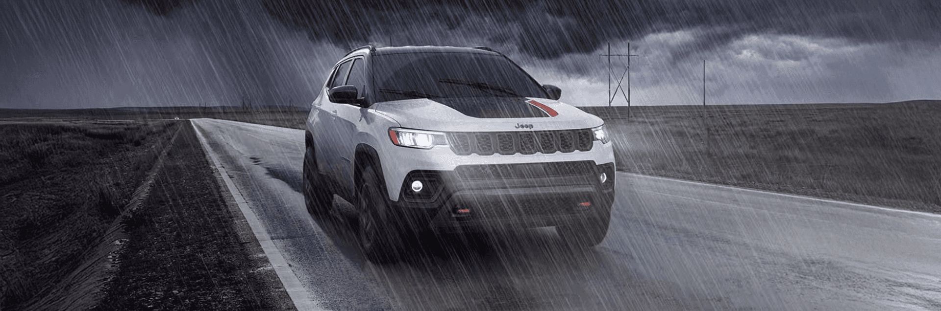 White 2026 Jeep Compass Trailhawk driving on a wet highway in heavy rain, showing black hood decal with red accent, LED lighting, and red tow hooks.