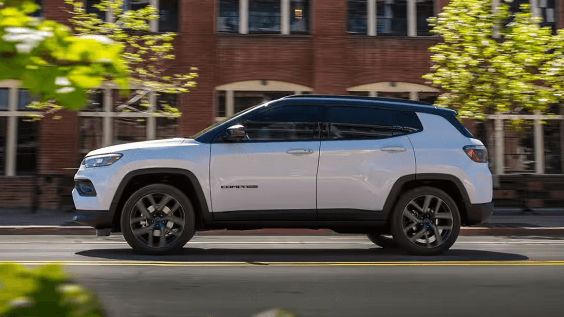 Side profile of a white Jeep Compass driving past brick buildings on a city street.