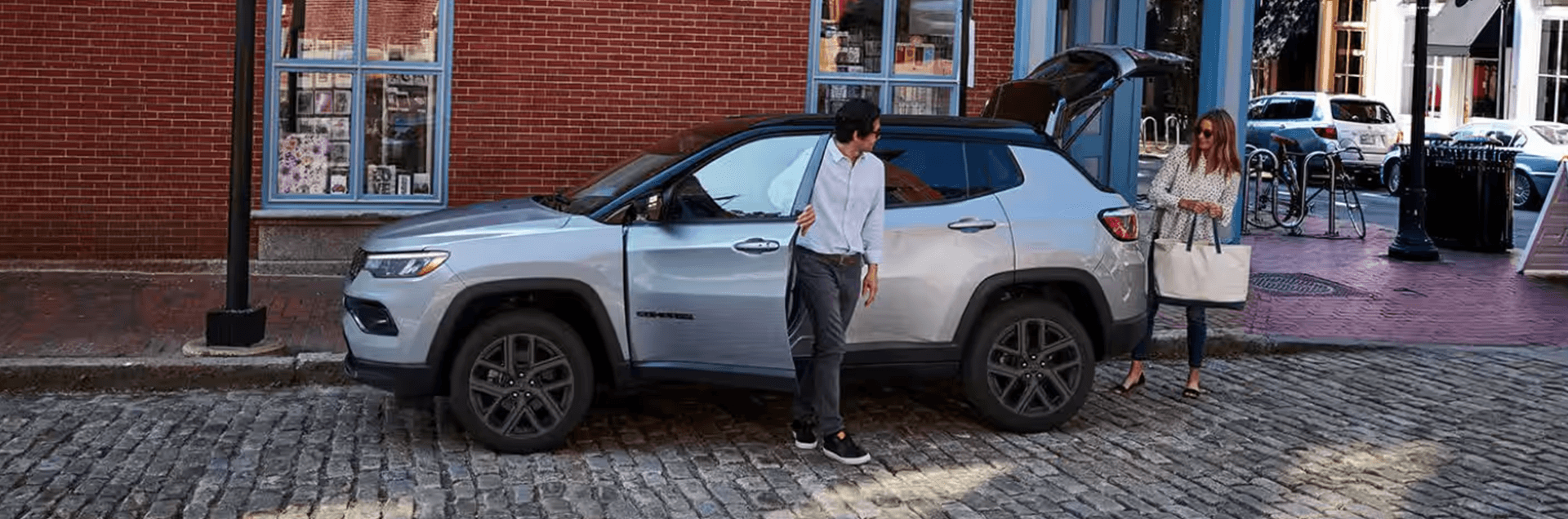 2026 Jeep Compass in silver parked on a cobblestone street with the rear liftgate open while two people load cargo near storefronts.