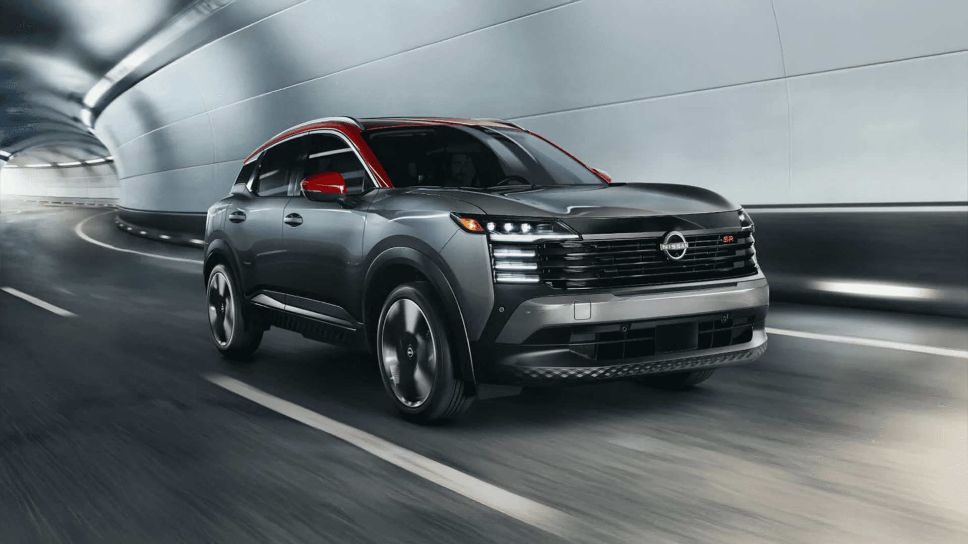 Gray 2026 Nissan Kicks driving through a tunnel, shown from a front three quarter angle with LED lighting, a black grille, and a red roof accent visible as the roadway blurs behind it