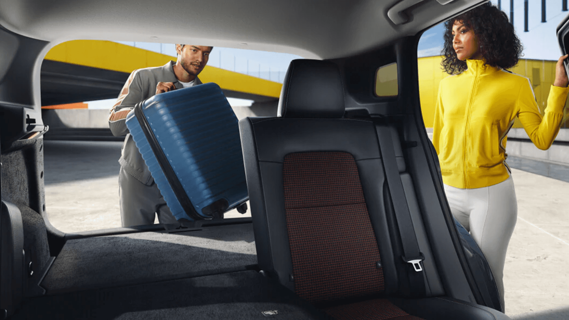 View from inside the rear of a 2026 Nissan Kicks showing the cargo area and rear seating while a person lifts a blue suitcase into the back and another person stands outside near the open hatch