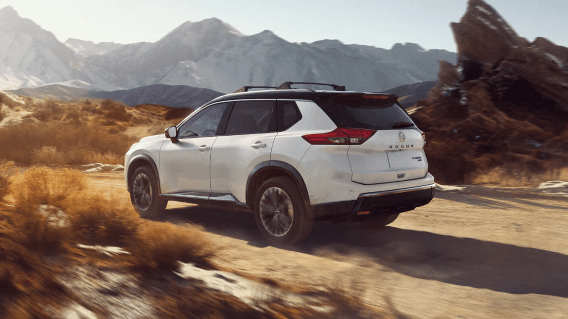 White Nissan Rogue rear three quarter view driving on a dirt road with mountains behind