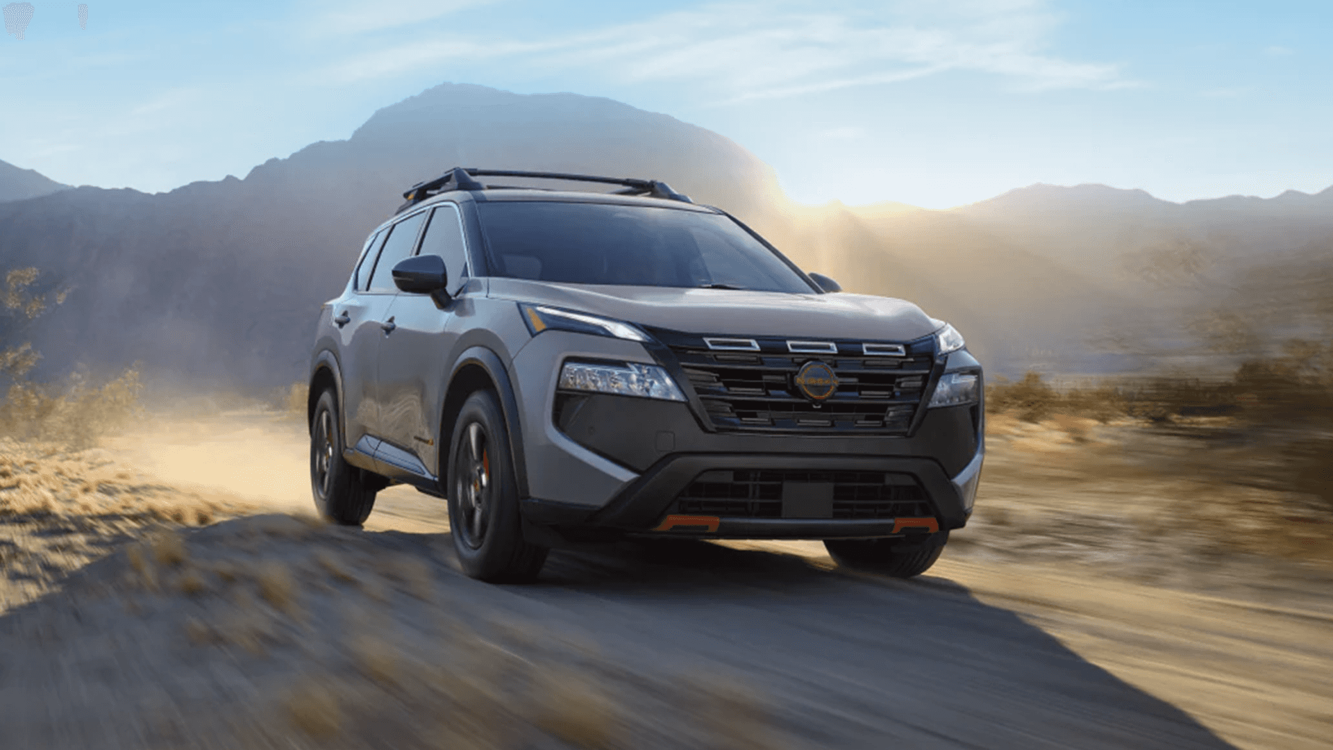 Gray Nissan Rogue driving on a dusty desert road with mountains in the background