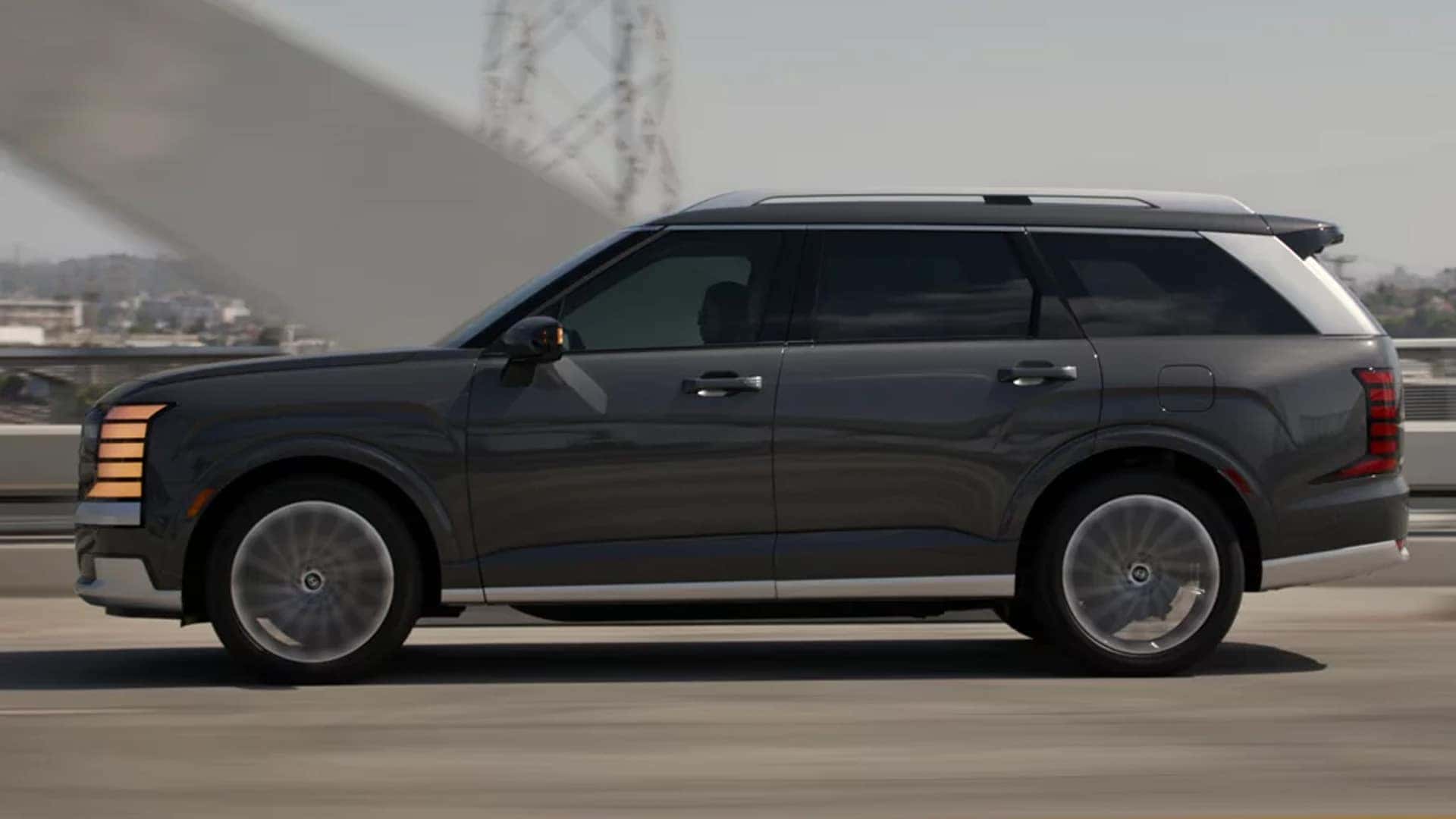 Side profile of the 2026 Hyundai Palisade Hybrid driving on a bridge, highlighting its three-row SUV design and hybrid performance