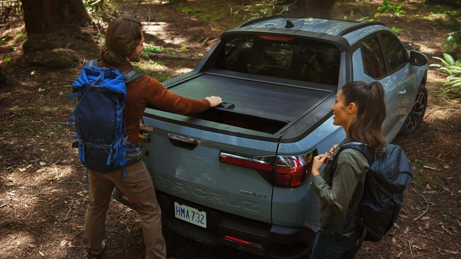 Rear view of a 2026 Hyundai Santa Cruz in a forest with the retractable bed cover partially open as hikers access gear from the truck bed.