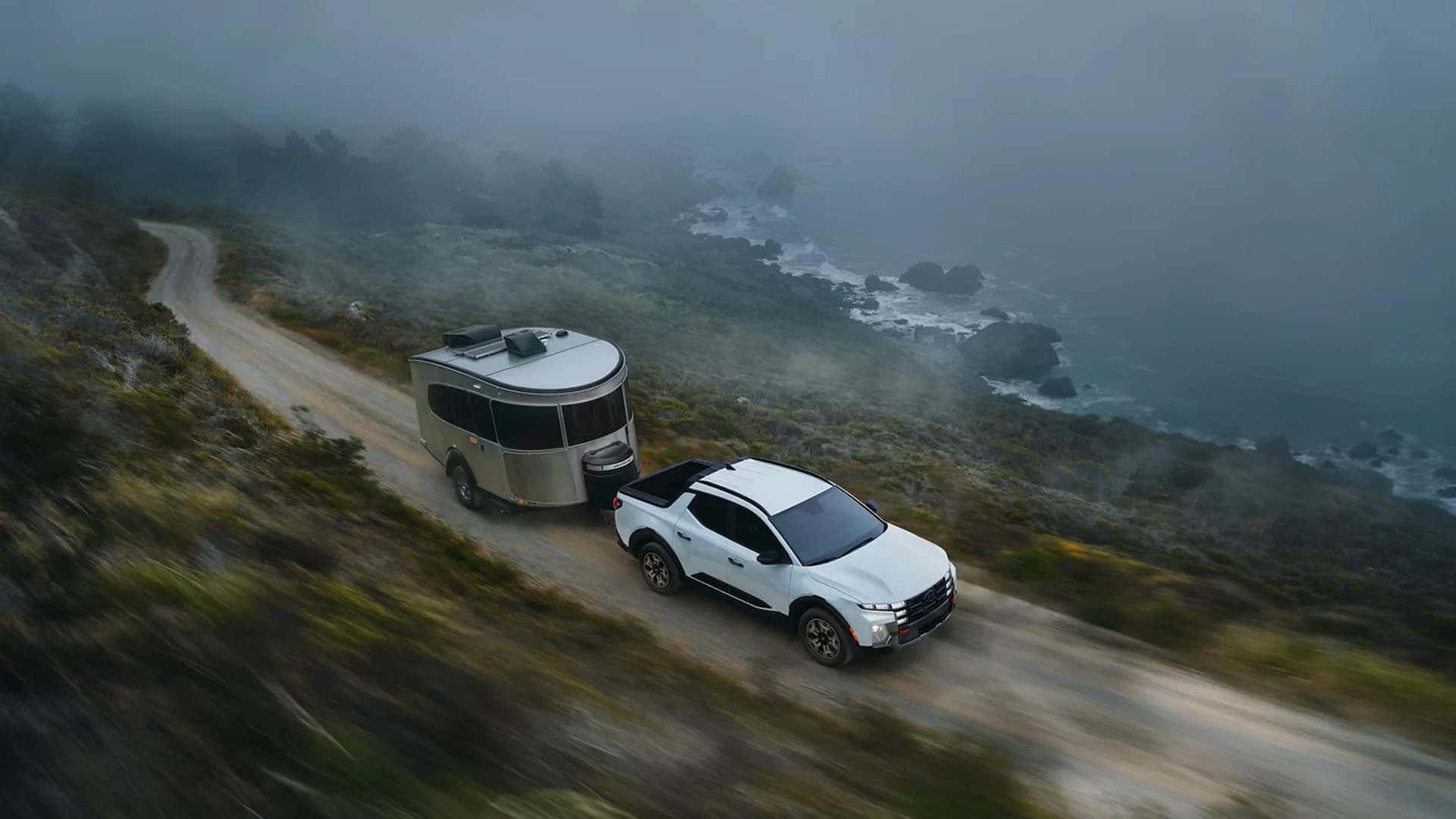 2026 Hyundai Santa Cruz towing a small travel trailer along a coastal road, demonstrating towing capability and utility performance