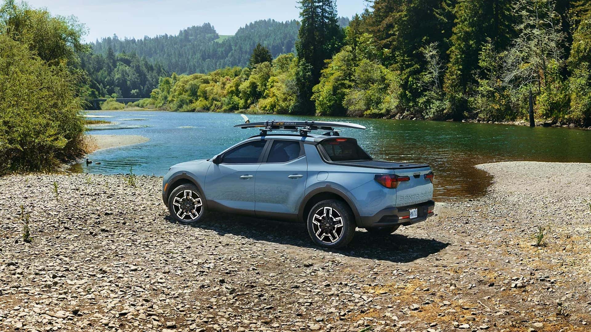 Blue 2026 Hyundai Santa Cruz parked on a rocky riverbank with roof mounted boards, showing rear three quarter view and outdoor styling.