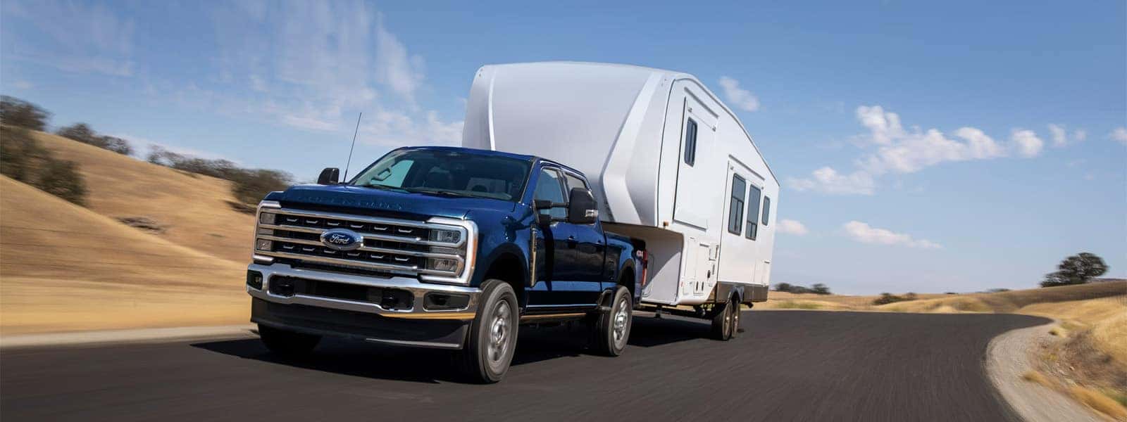 2026 Ford F-250 Super Duty towing a large fifth wheel camper on a highway through rolling hills