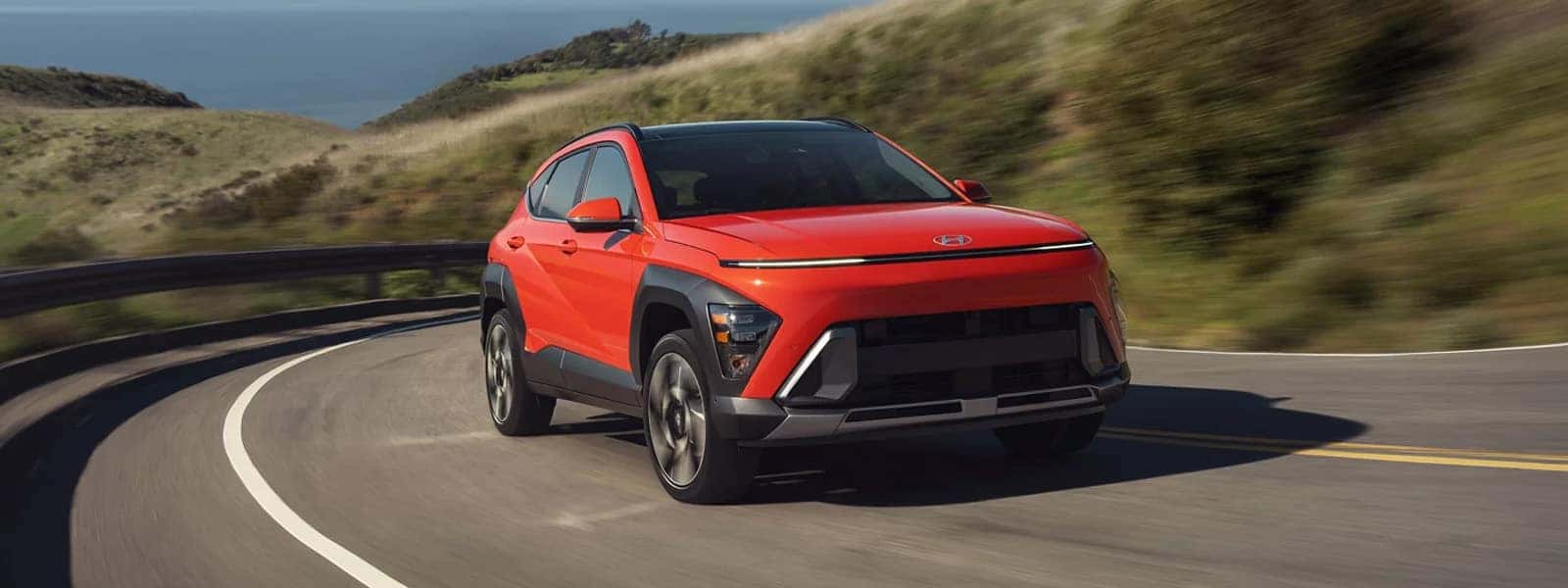 Red 2026 Hyundai Kona driving on a winding coastal road, showing bold front grille, LED lighting strip, and compact SUV proportions.