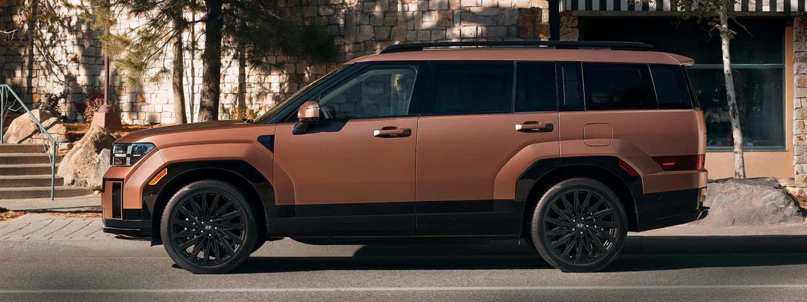 Bronze 2026 Hyundai Santa Fe parked curbside, showing boxy exterior design, black roof rails, dark alloy wheels, and upright midsize SUV proportions.