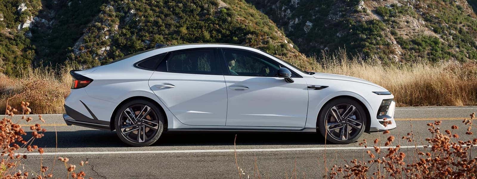 2026 Hyundai Sonata driving on a scenic road, showing sleek sedan profile, low stance, and modern exterior design
