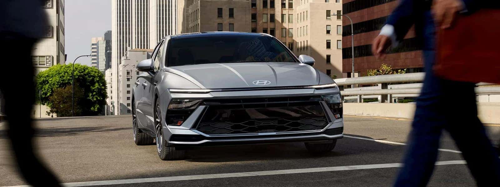 Front view of a 2026 Hyundai Sonata Hybrid driving through a downtown city street, highlighting wide grille design and slim LED daytime running lights.