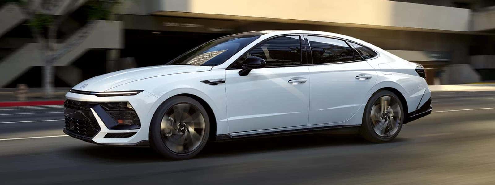 2026 Hyundai Sonata Hybrid driving through a city street, showcasing sleek midsize sedan styling, aerodynamic profile, and modern LED lighting design