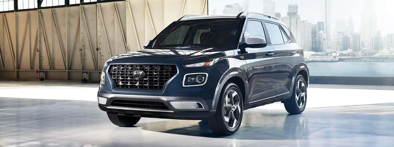 2026 Hyundai Venue parked inside a modern industrial space with city skyline in the background, showcasing bold grille and compact SUV stance.
