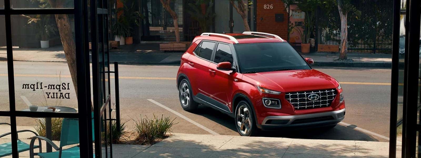 Red 2026 Hyundai Venue parked on a city street, viewed from a cafe, highlighting compact SUV styling and roof rails