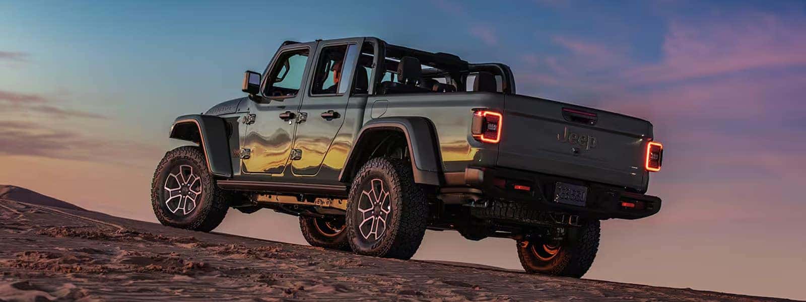 Gray 2026 Jeep Gladiator pickup on sand at sunset with open roof, chunky off-road tires, and LED taillights.