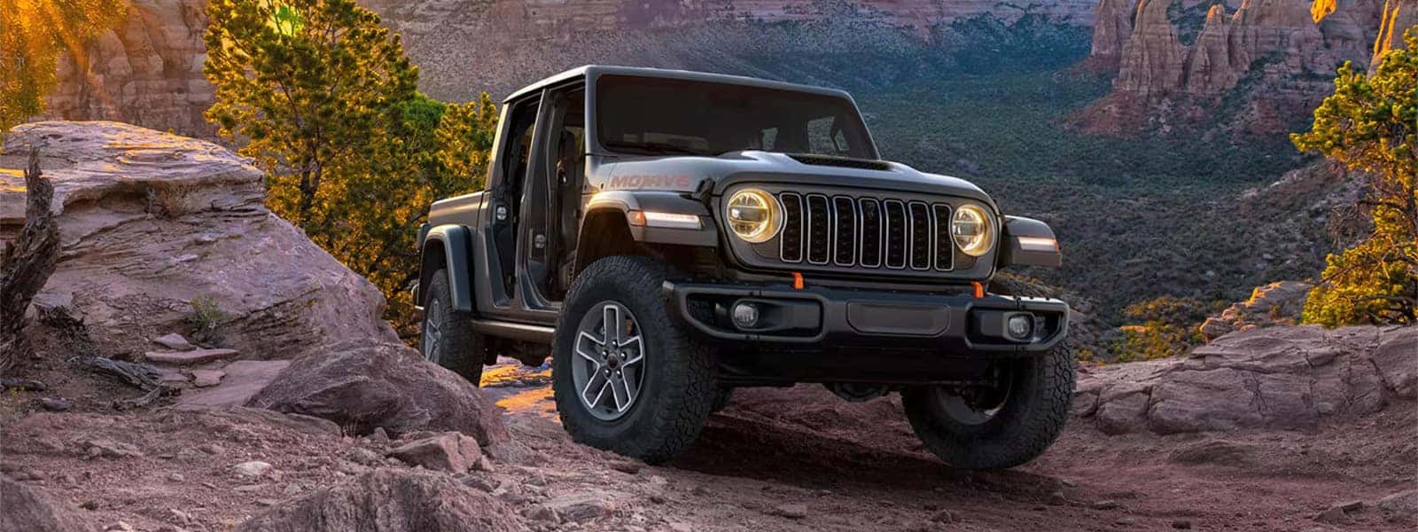 2026 Jeep Gladiator Mojave on a rocky trail at sunset, showing open-air pickup design and rugged front stance