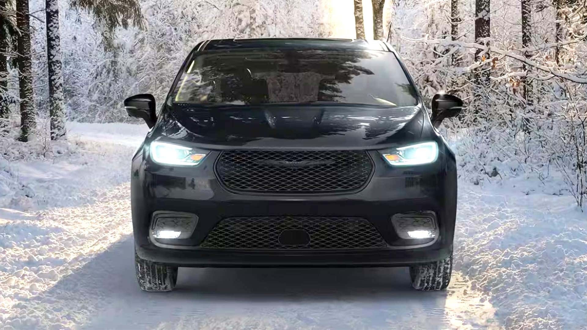 Front view of 2026 Chrysler Pacifica driving on a snowy road with LED headlights and mesh grille illuminated