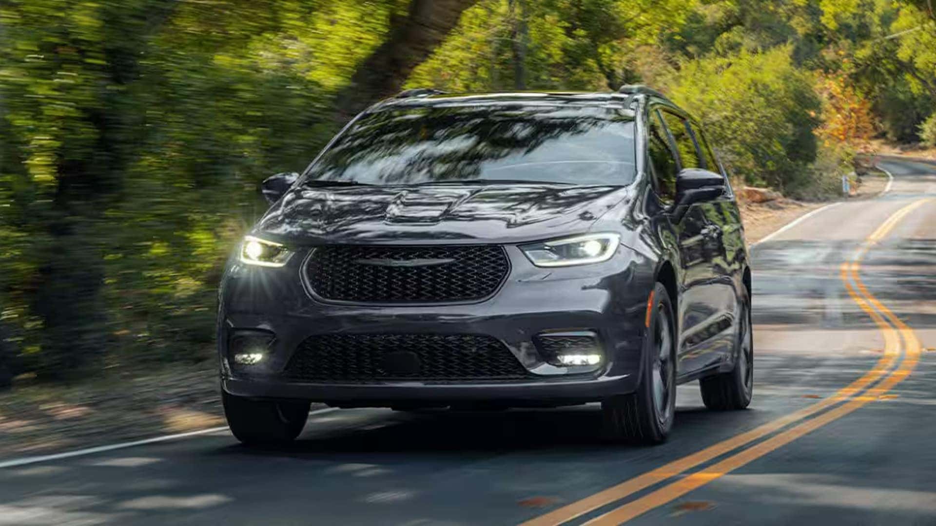 2026 Chrysler Pacifica driving on a winding road through trees, showing the front grille and LED headlights
