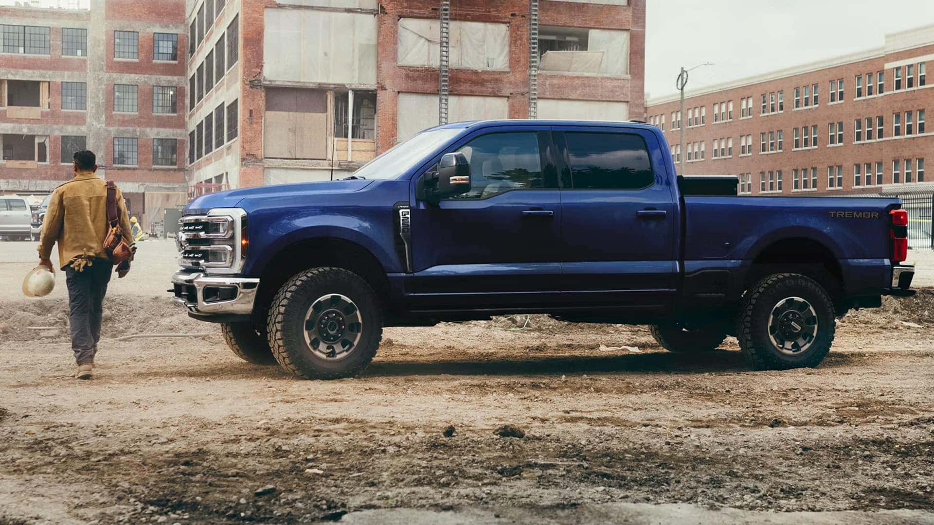 Side profile of a blue 2026 Ford F-250 Super Duty on a construction site, highlighting crew cab configuration, high ground clearance, and Tremor off-road package styling