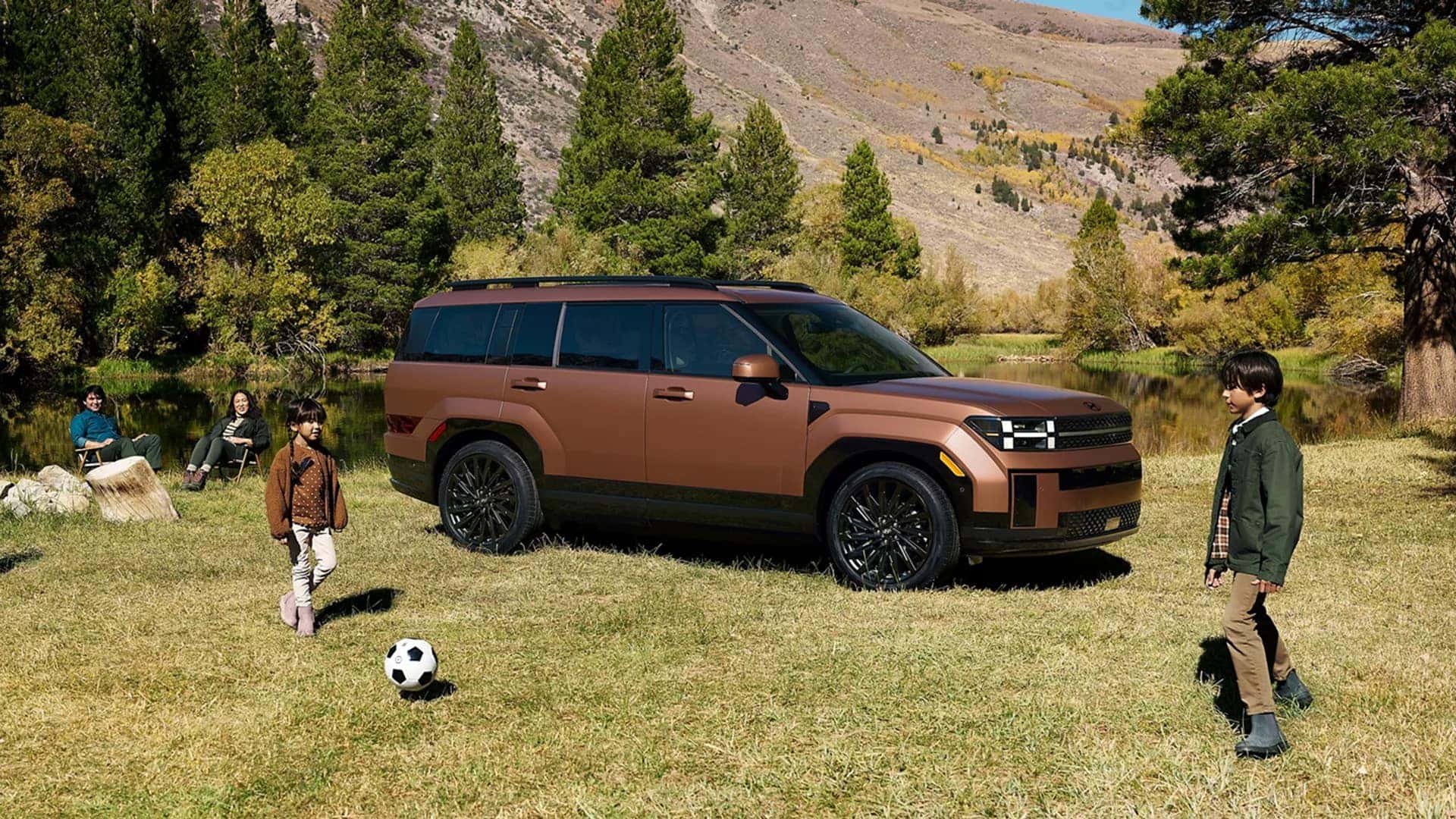 Bronze 2026 Hyundai Santa Fe parked on grass near trees and mountains, highlighting squared profile, long roofline, and rugged exterior styling.