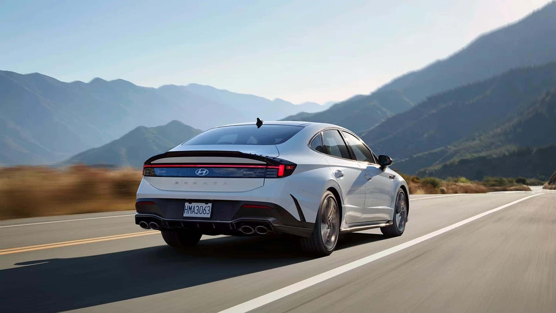 Rear three quarter view of a 2026 Hyundai Sonata Hybrid on a scenic highway, showing full width LED tail light bar and sleek sedan profile.