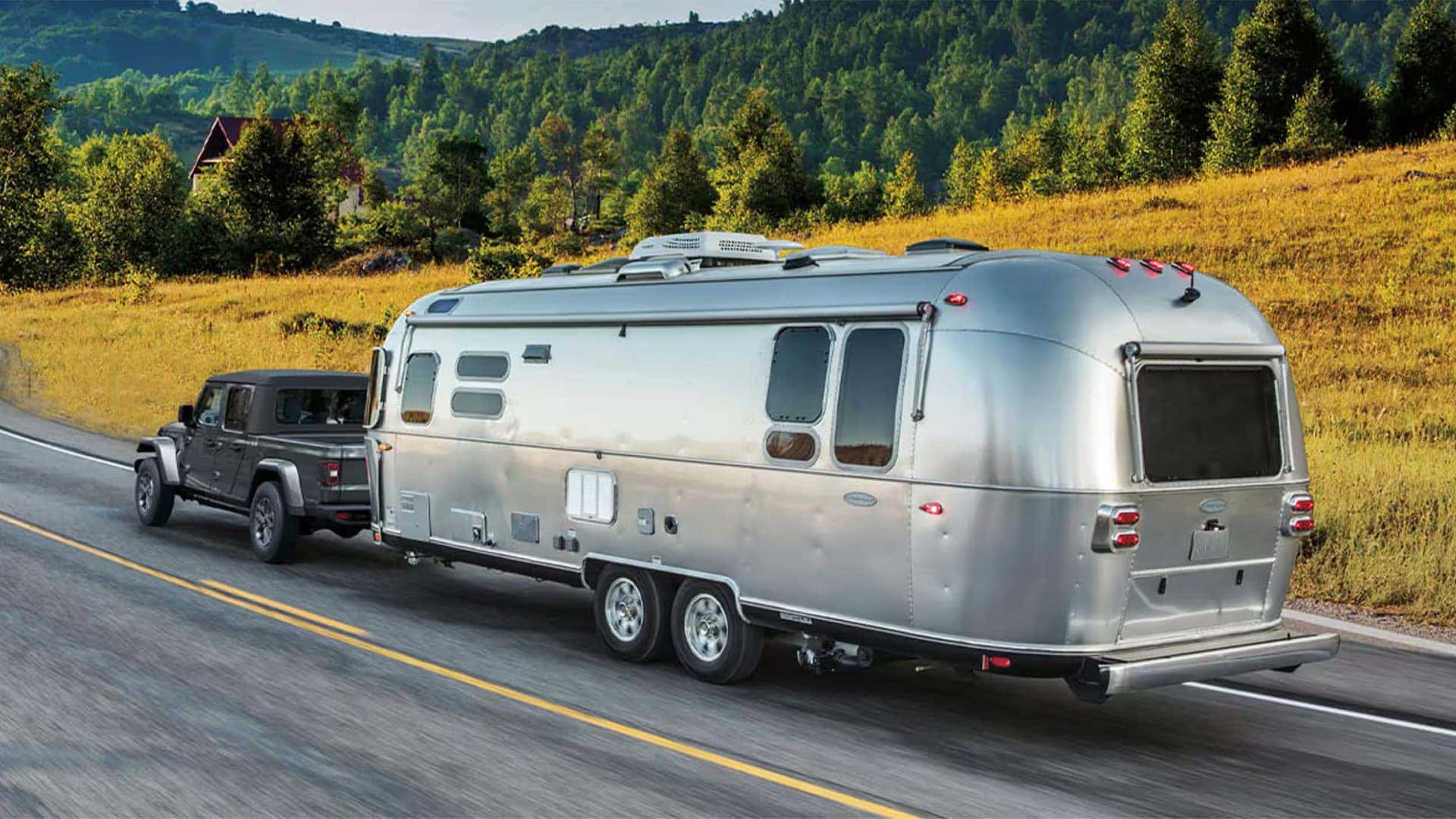 2026 Jeep Gladiator towing a silver travel trailer on a scenic highway through rolling hills