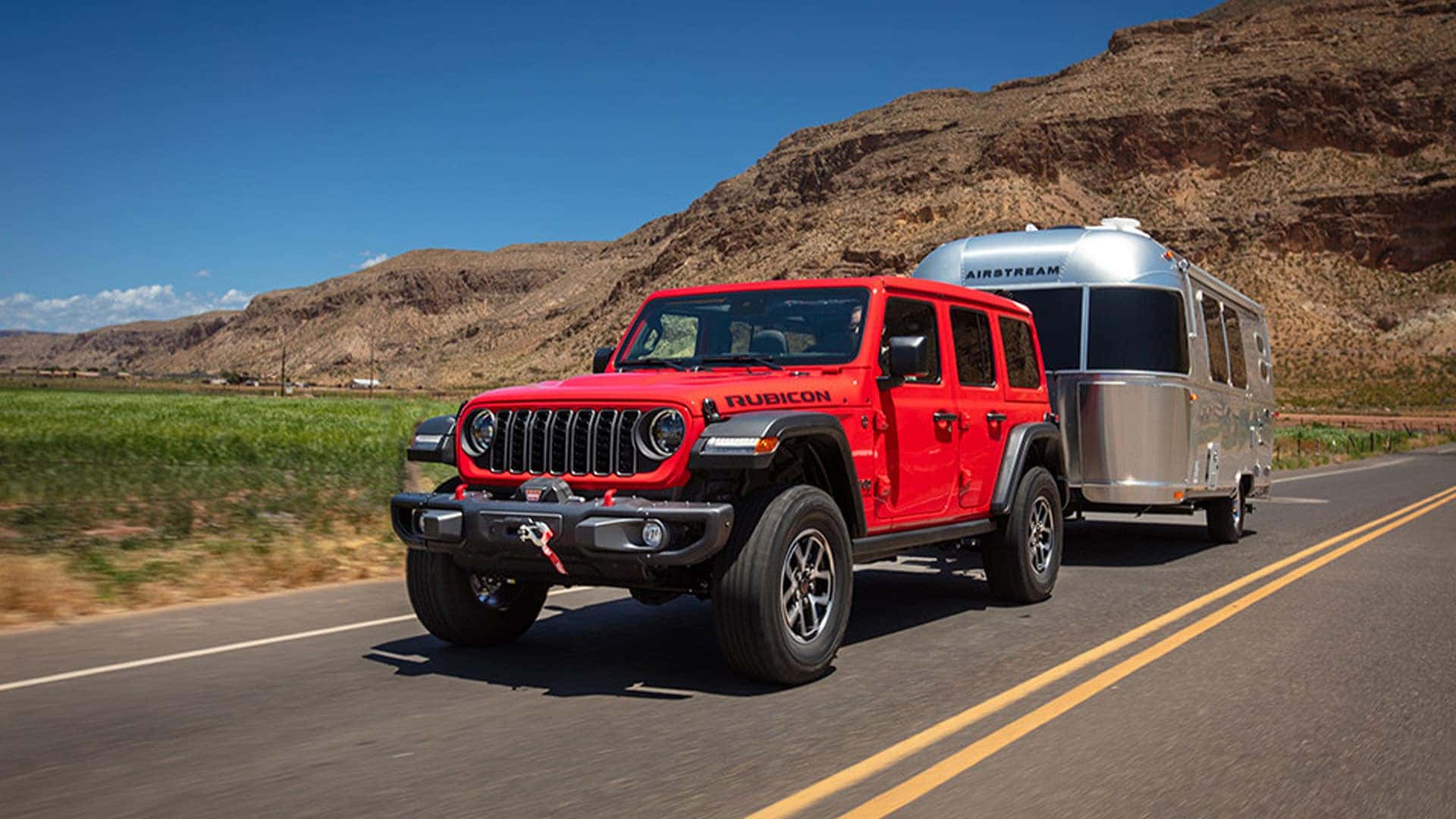 Red 2026 Jeep Wrangler Rubicon towing a silver Airstream travel trailer on a desert highway