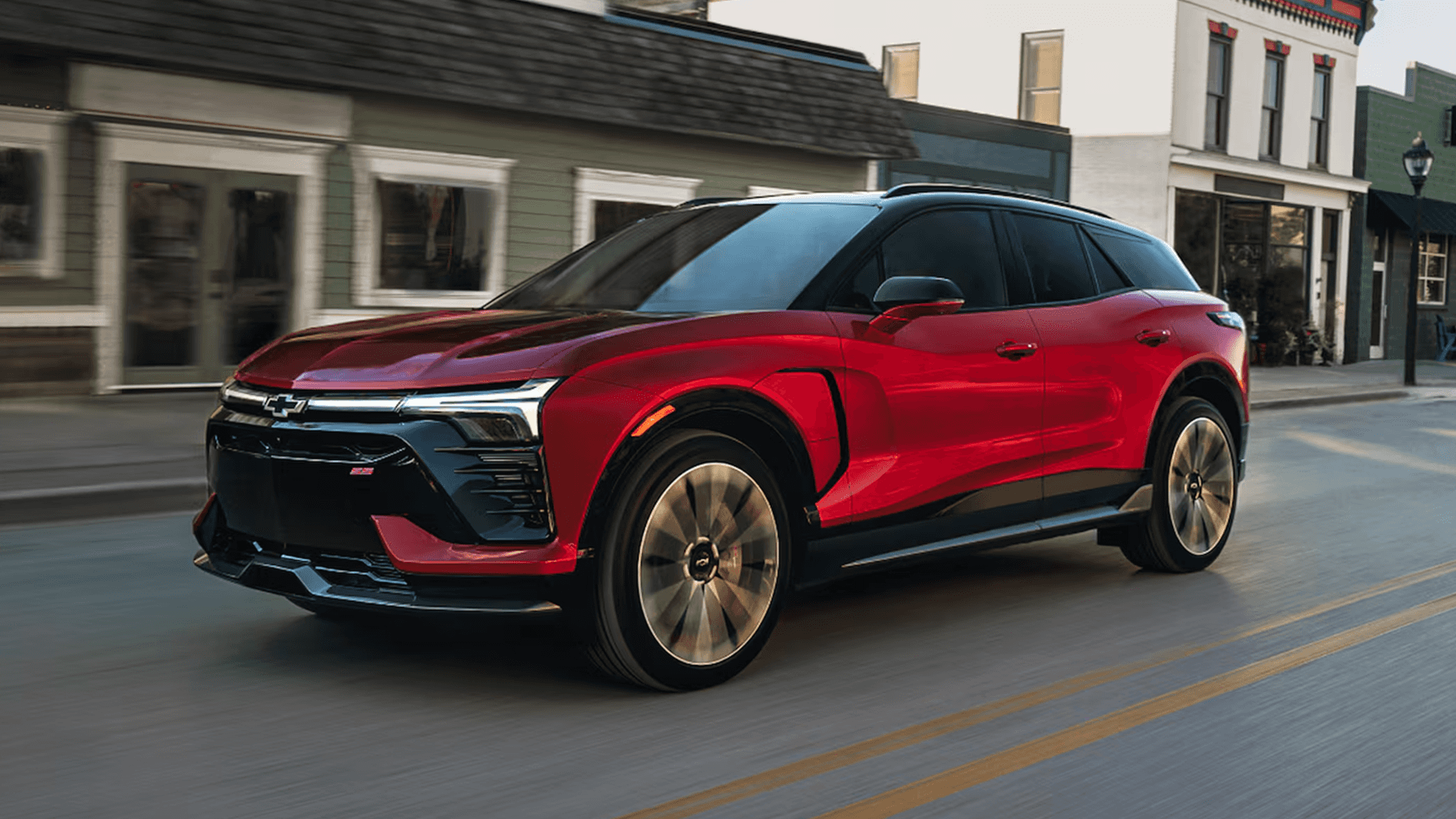 Red 2026 Chevy Blazer EV driving through a small town street with storefronts, showing side profile and motion blur