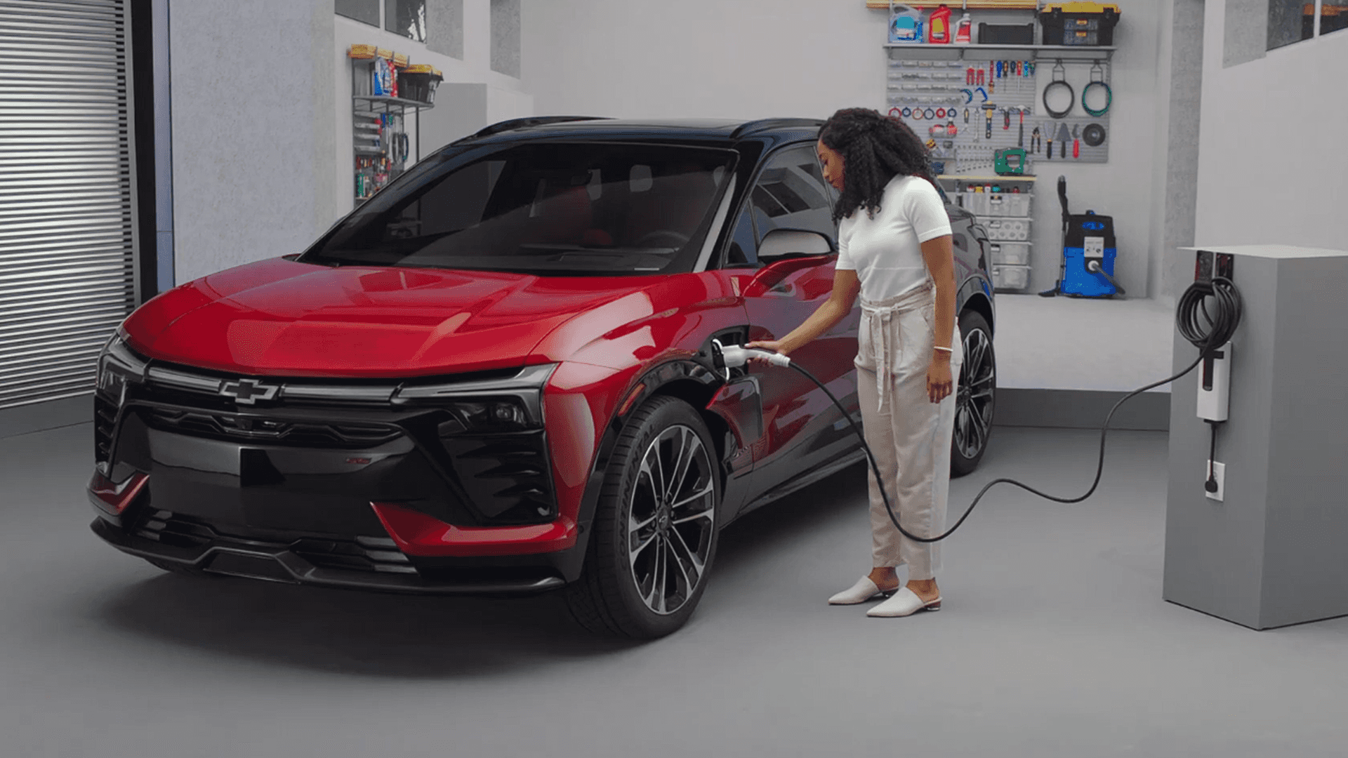 Woman plugs charging cable into red Blazer EV in garage, home charger on pedestal and tools on wall