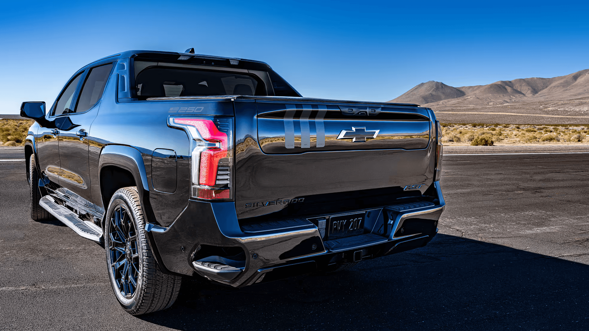 Rear three quarter view of a dark Silverado EV on open pavement with mountains, showing tail lamps and badging