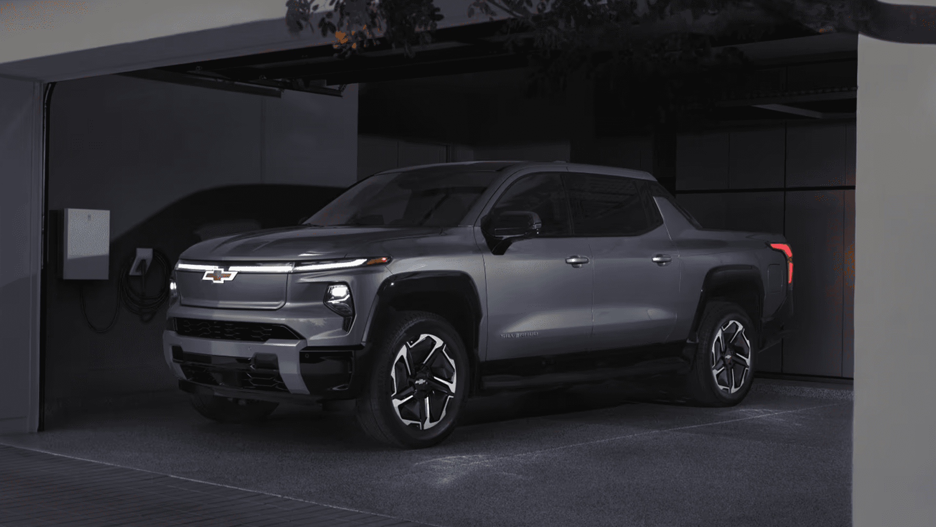 Gray Chevy Silverado EV parked in a modern driveway beside a home charging station, shown from a front three quarter angle.