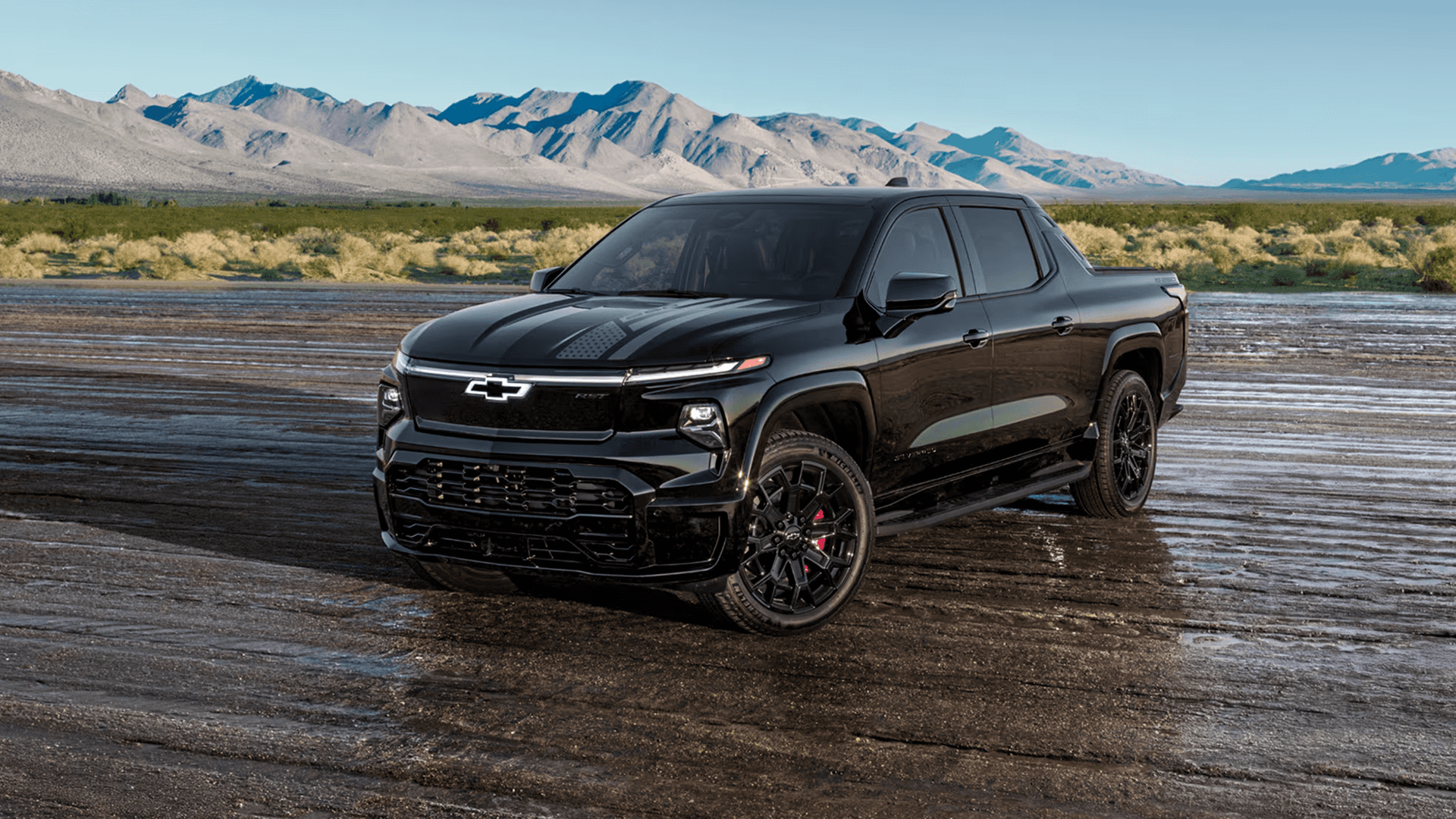 Black Chevy Silverado EV parked on a wide desert plain with mountains in the background, shown from a front three quarter view.