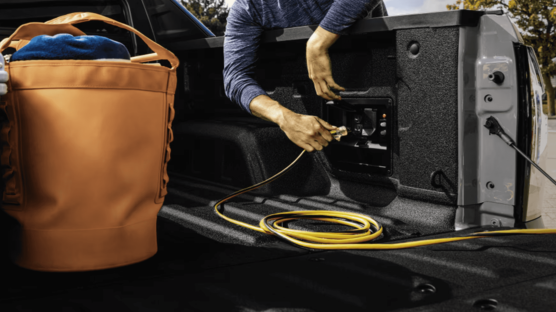 Person plugging a power cord into a bed-mounted outlet panel inside the Silverado EV cargo box next to a tote bag.