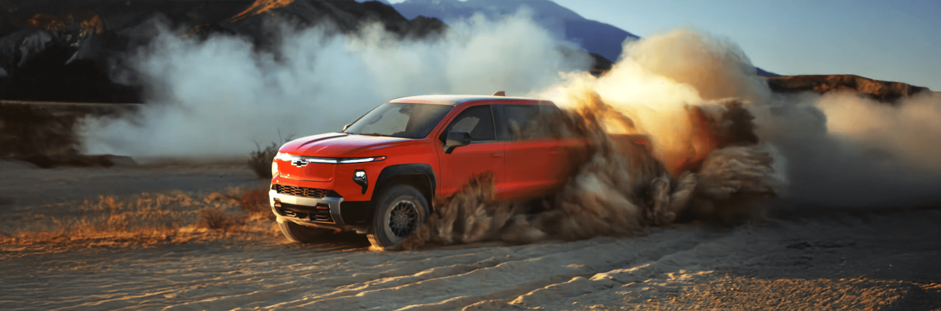 Red Chevy Silverado EV powers through sand, kicking up dust clouds with mountains in the distance.