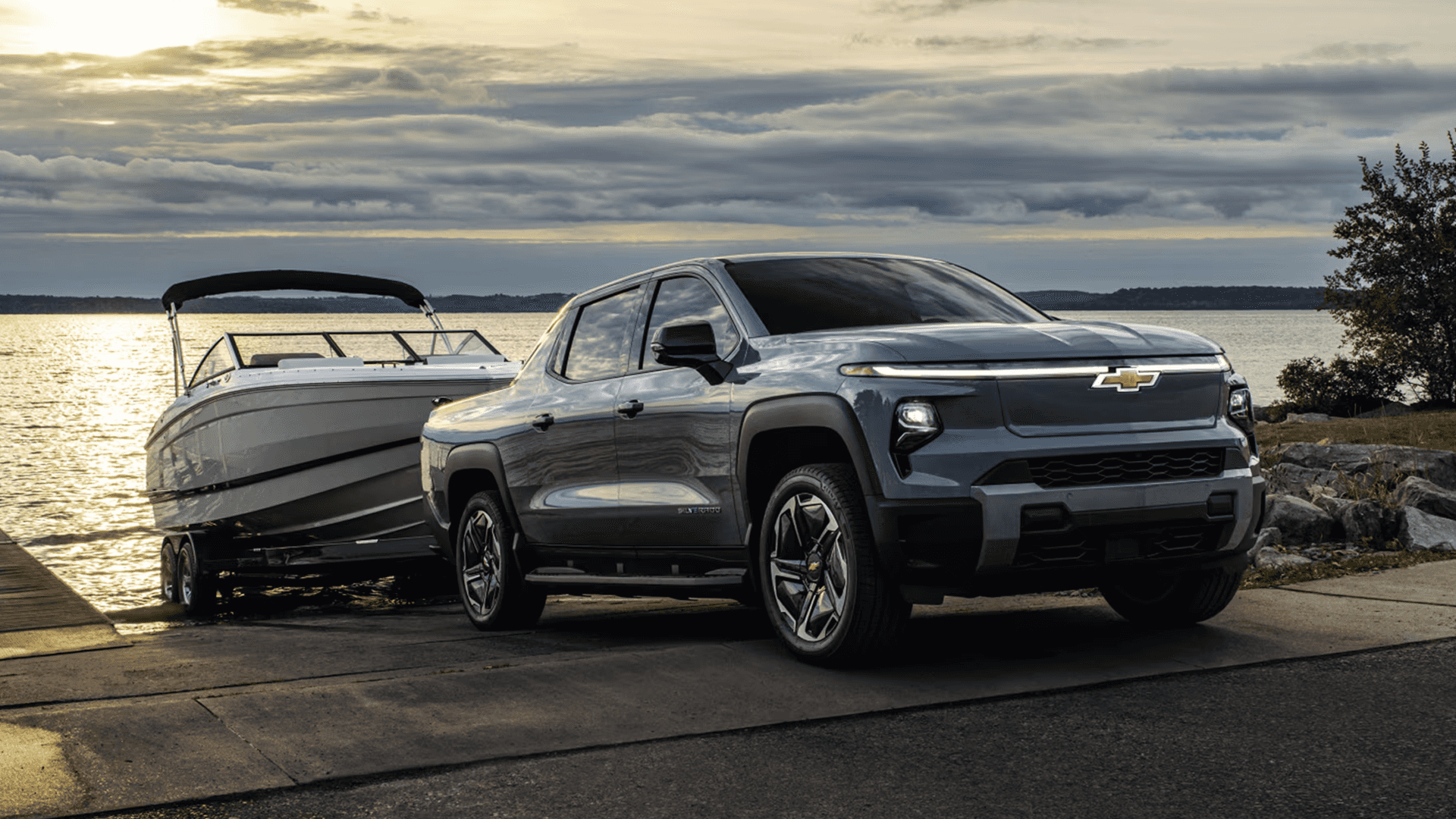 Gray 2026 Chevy Silverado EV towing a boat on a ramp beside a lake at sunset under cloudy skies