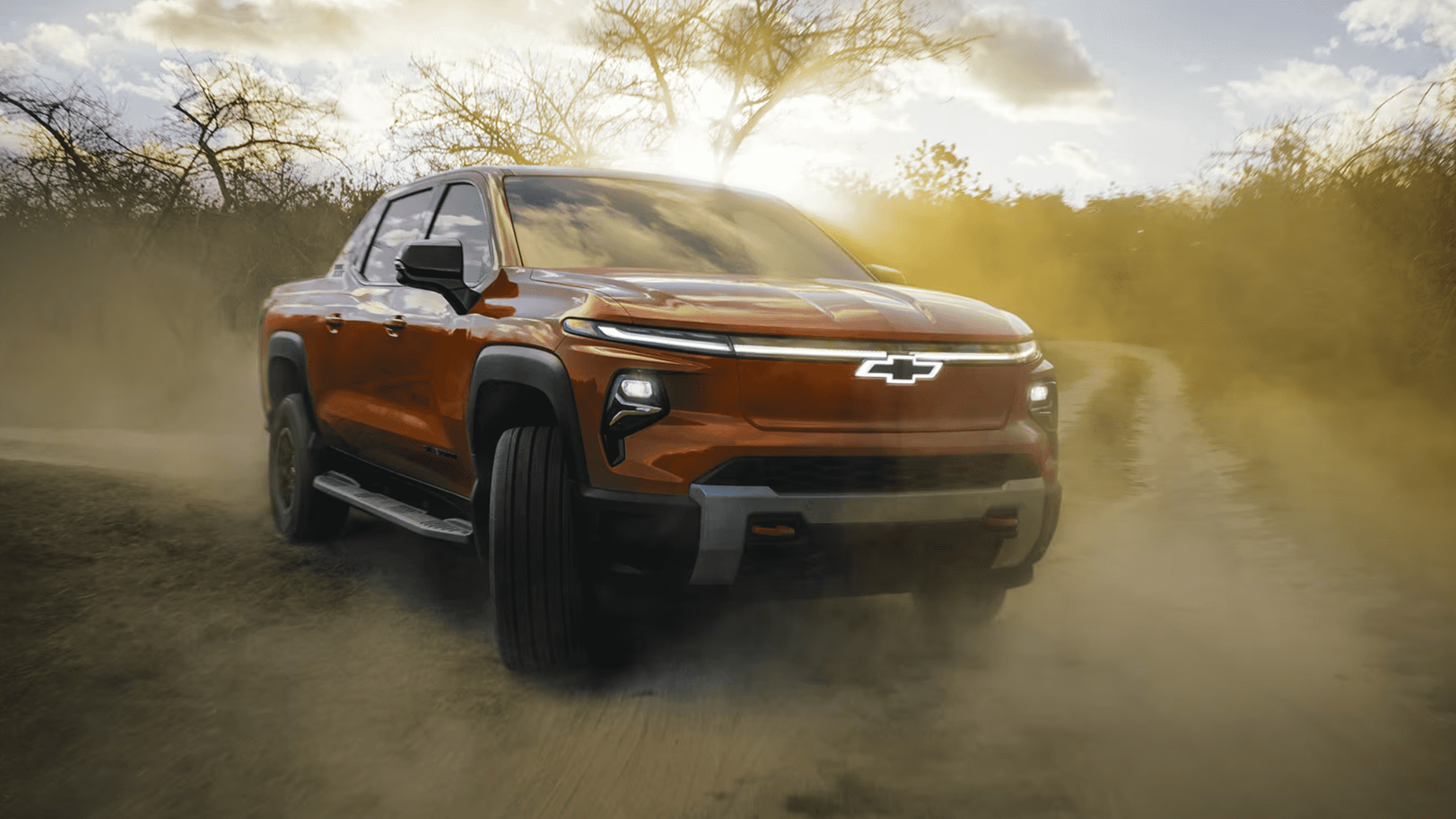 Orange 2026 Chevy Silverado EV kicking up dust on a winding dirt road with sunlight through trees