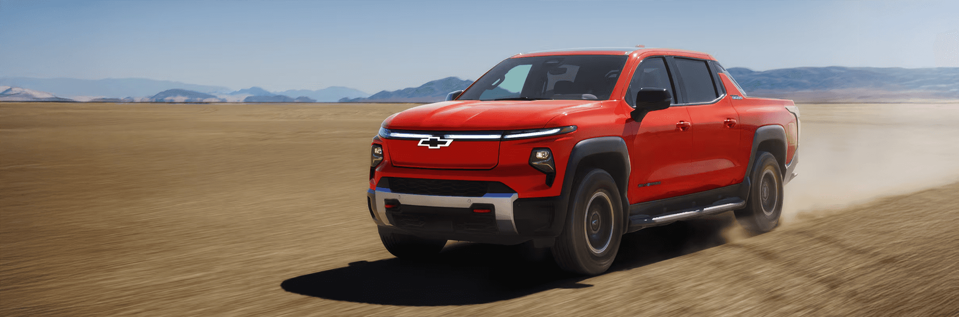 Orange 2026 Chevy Silverado EV driving across a wide desert landscape with mountains in the distance