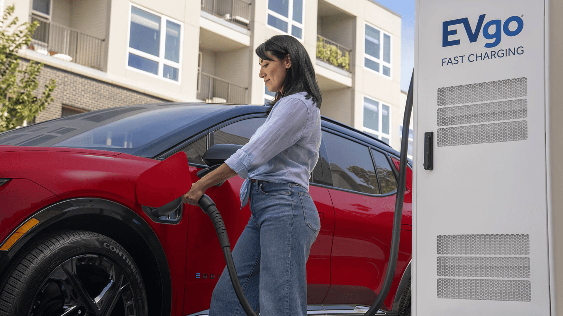 Driver plugs red Chevy Equinox EV into EVgo fast charger outside apartment building in daylight