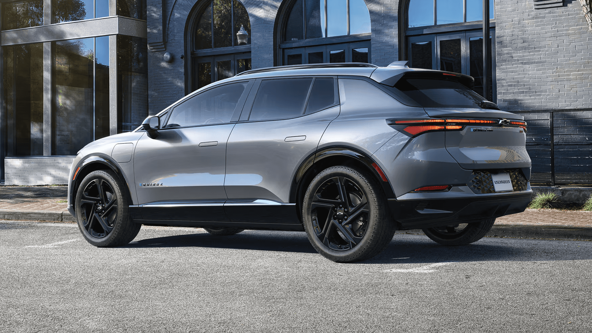 Gray Chevy Equinox EV rear three quarter view outside brick building, featuring LED tail lamp bar and black wheels.