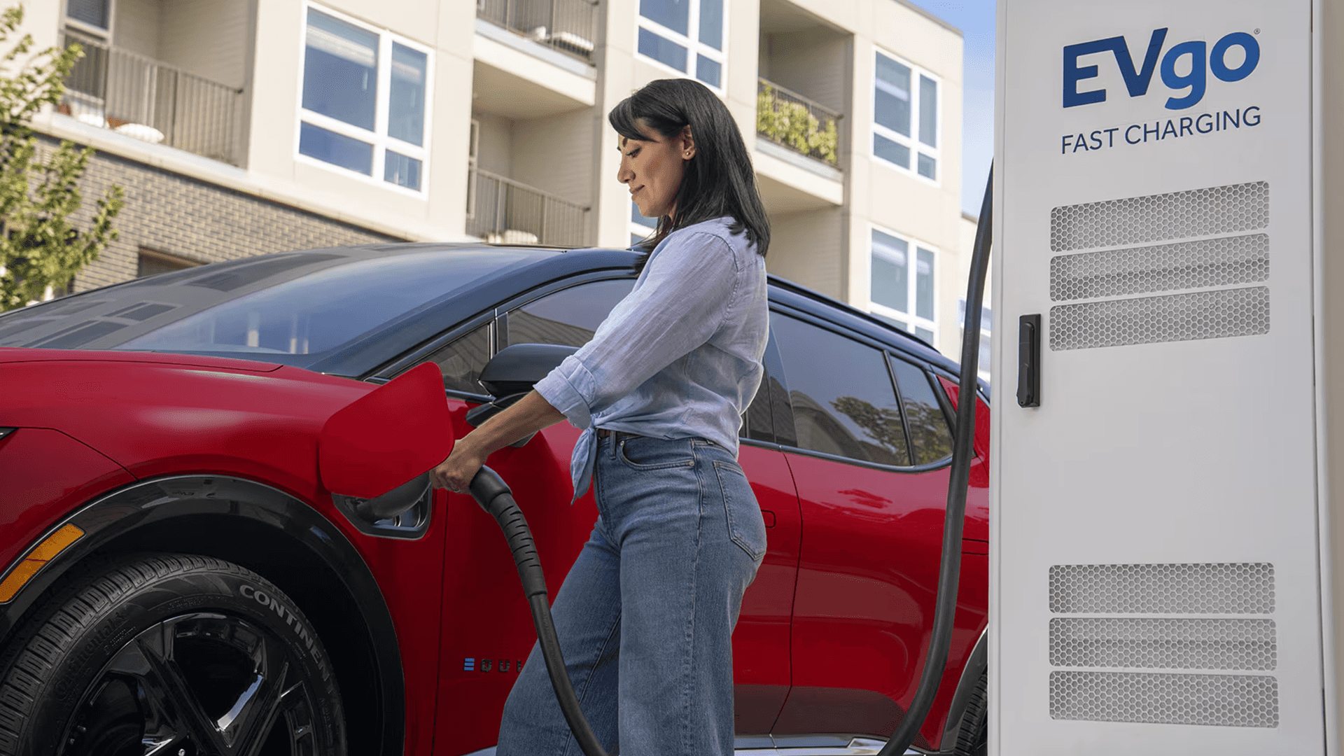 Driver plugs a red Chevy Equinox EV into an EVgo fast charging station outside modern apartment buildings.