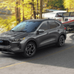 Gray 2026 Ford Escape towing a boat on a curving wooded road, showing towing capability and stability.