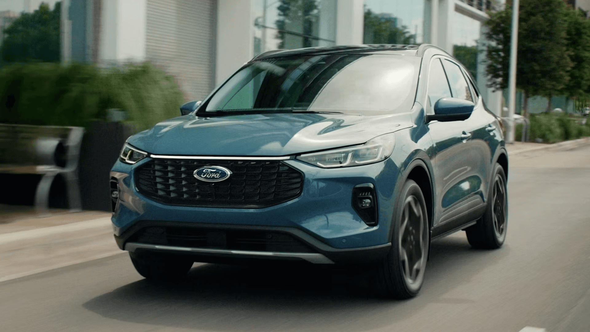 Blue 2026 Ford Escape driving past modern buildings, showing grille, LED lights, and sleek profile