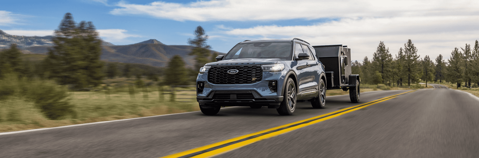 2026 Ford Explorer ST towing a black trailer on an open two-lane road with forest scenery