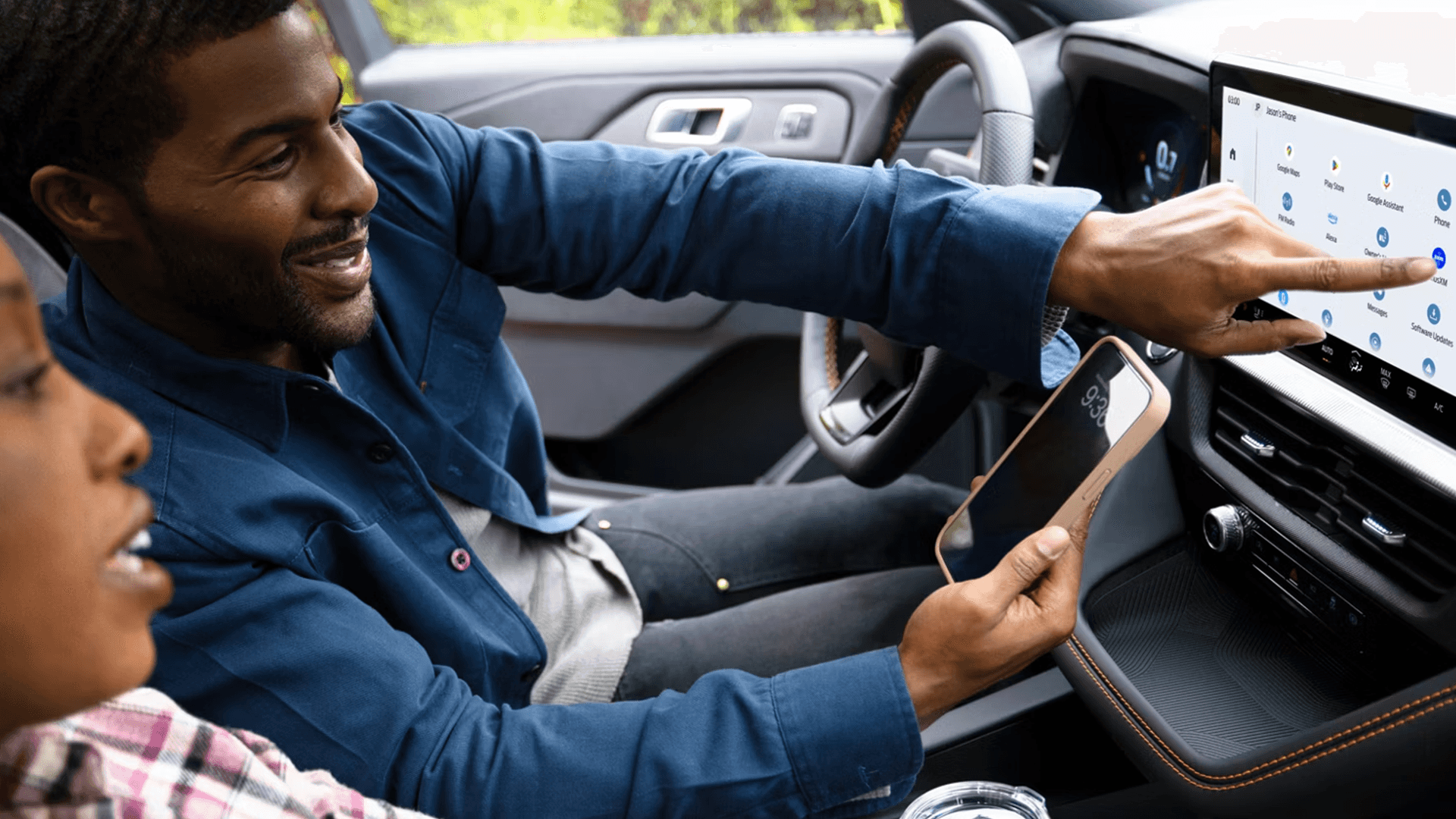 Driver points to Explorer touchscreen apps while holding a smartphone, demonstrating connected infotainment features