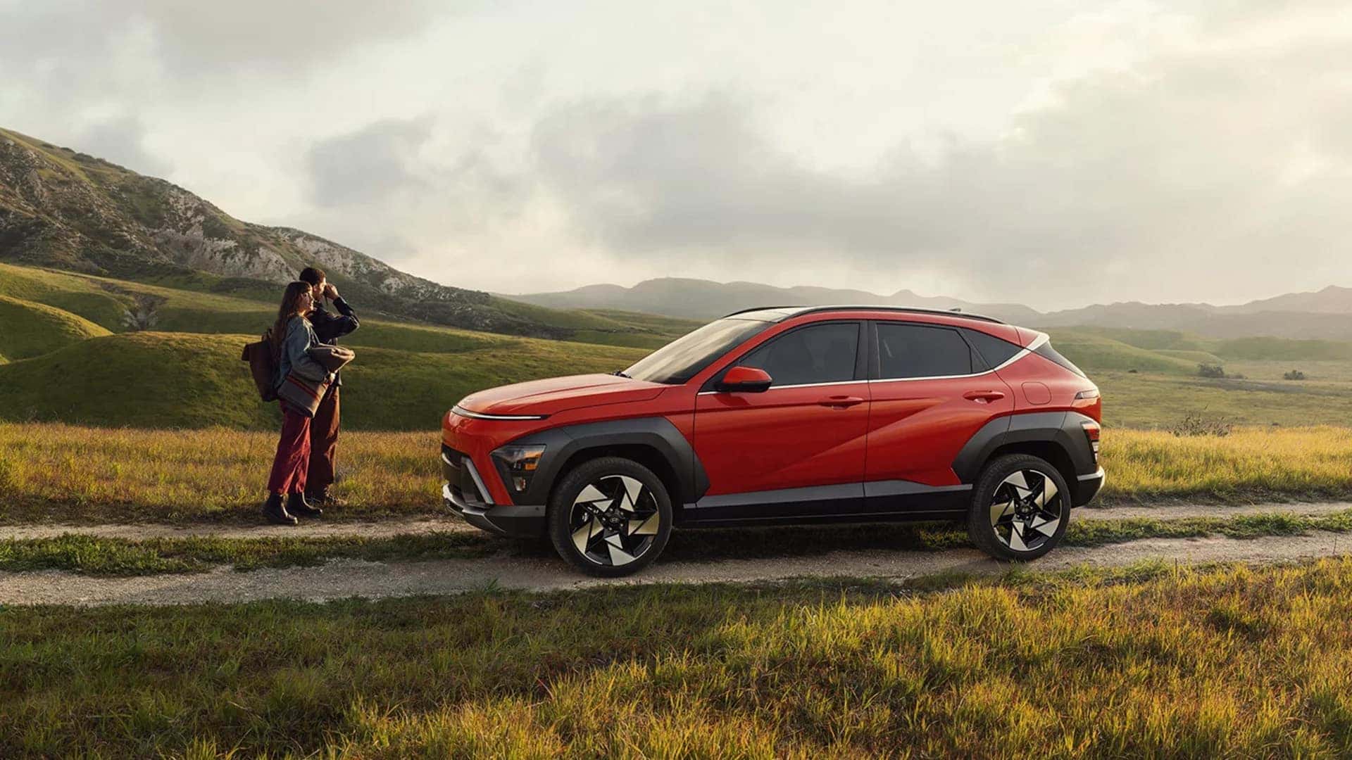 Side profile of a red 2026 Hyundai Kona parked in an open landscape, showing wheel design, body cladding, and compact SUV stance.