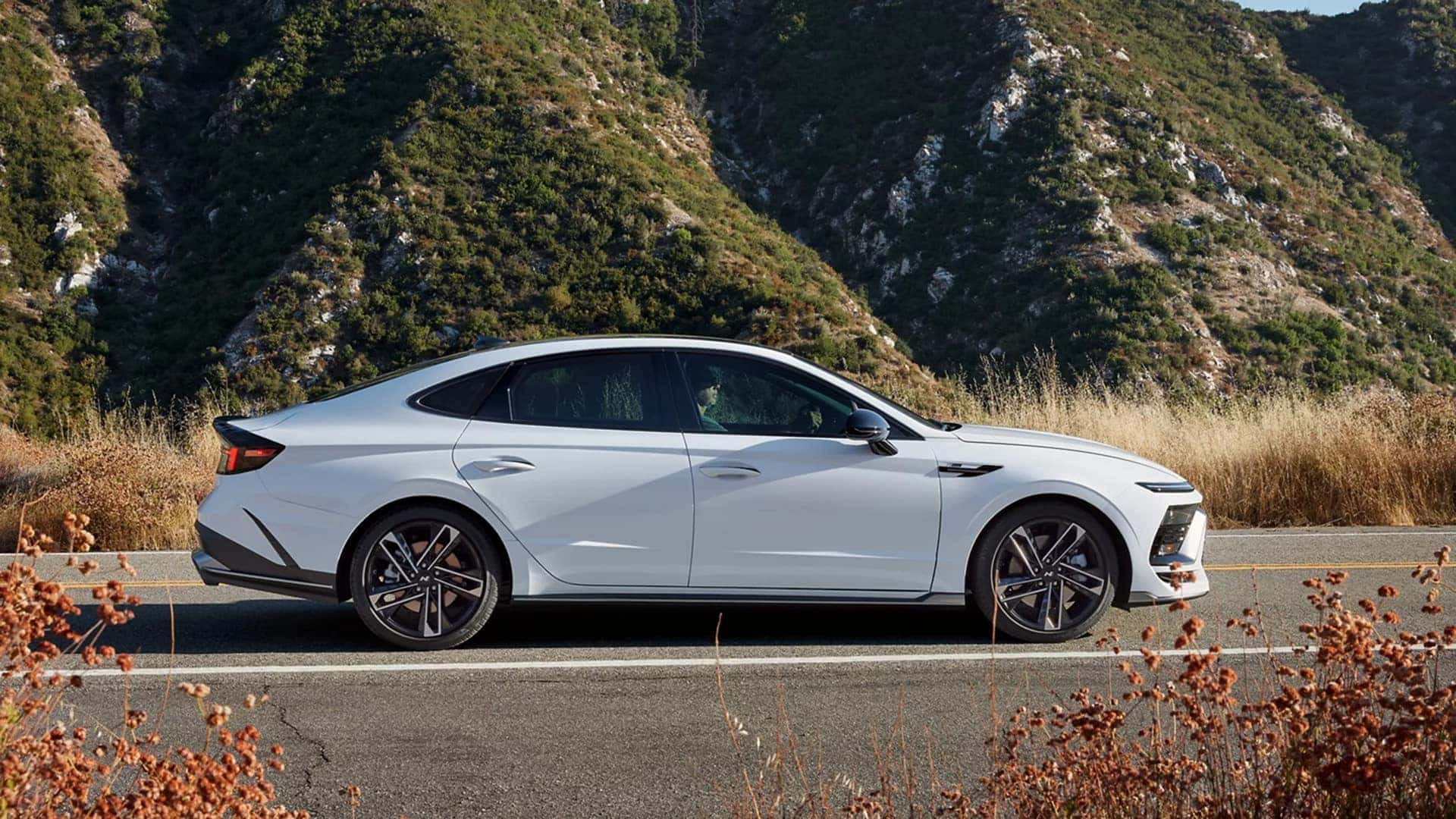 Side profile of a 2026 Hyundai Sonata Hybrid parked on a mountain road, showcasing aerodynamic roofline and alloy wheel design.