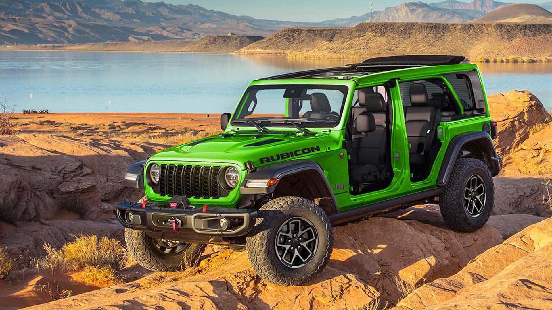 Green 2026 Jeep Wrangler Rubicon with doors removed on rocky shoreline, showing open-air cabin and off-road tires.