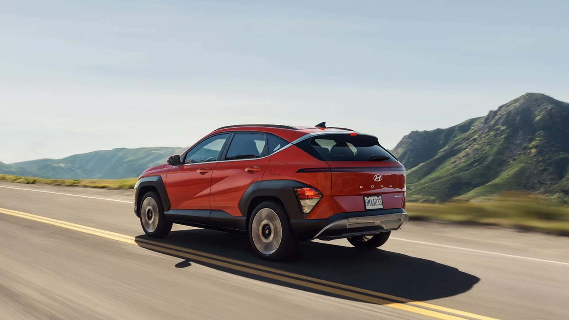Rear three quarter view of a red 2026 Hyundai Kona driving on an open highway, emphasizing compact SUV handling, smooth ride, and aerodynamic design
