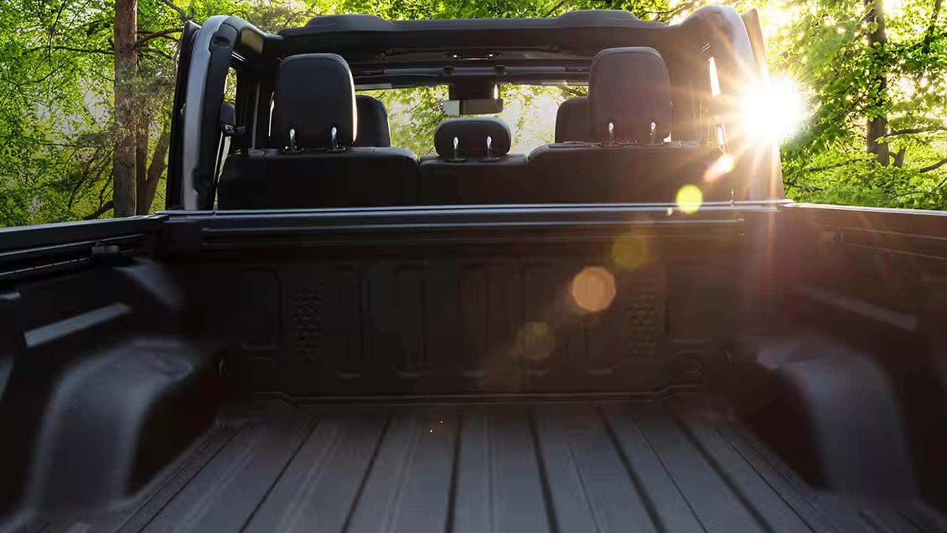 View from inside the 2026 Jeep Gladiator truck bed looking toward the cab with open roof and sunlight through trees.